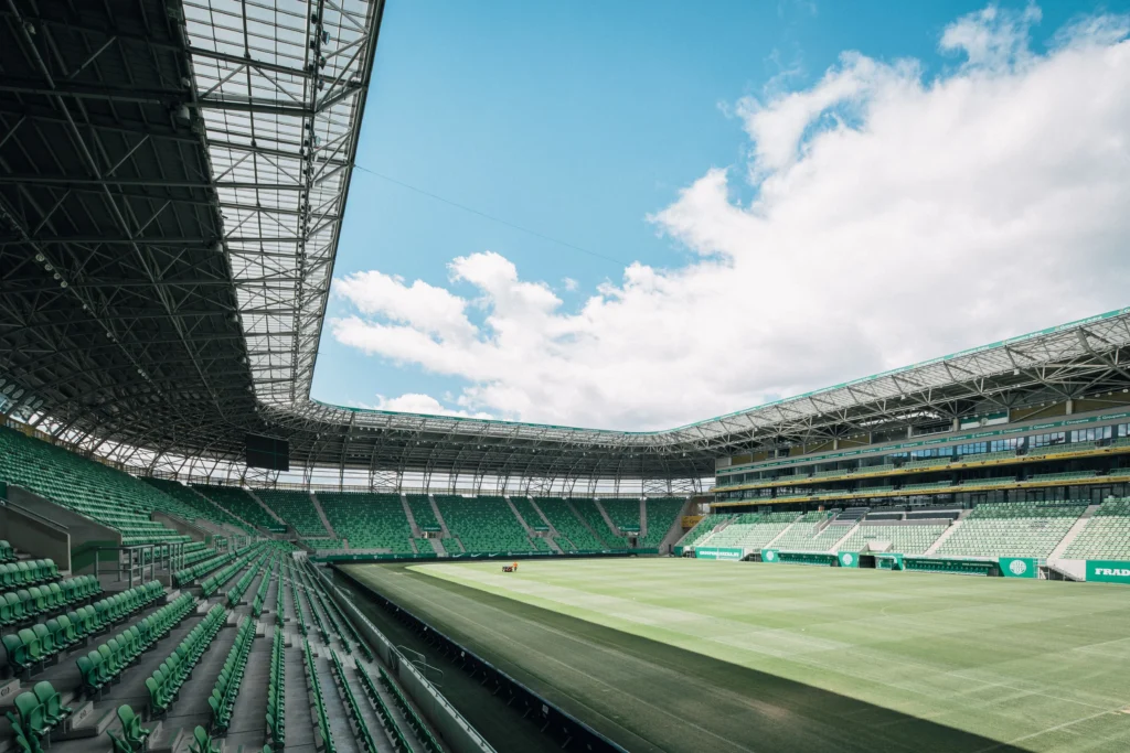Stadium interior with rows of green seats, steel framework, and a bright blue sky above the open roof.