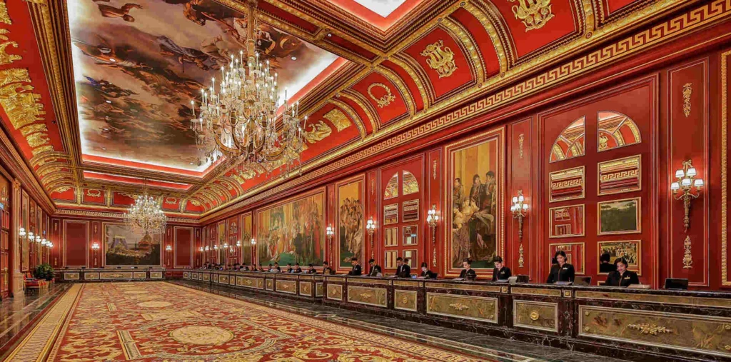 Opulent red-and-gold ceremonial hall with chandeliers and long desks along the wall, staff seated at intervals.