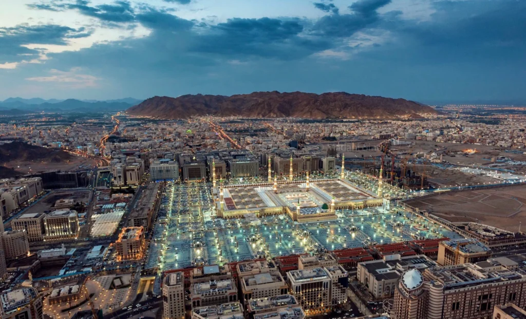 Aerial view of Mecca's Grand Mosque at dusk, with illuminated courtyards and surrounding city against distant mountains.