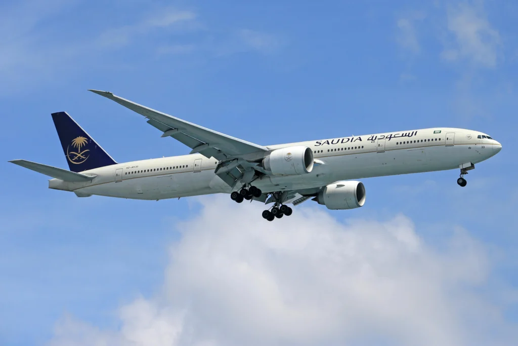Saudi Arabian Airlines aircraft in flight, side view with landing gear down against a blue sky.