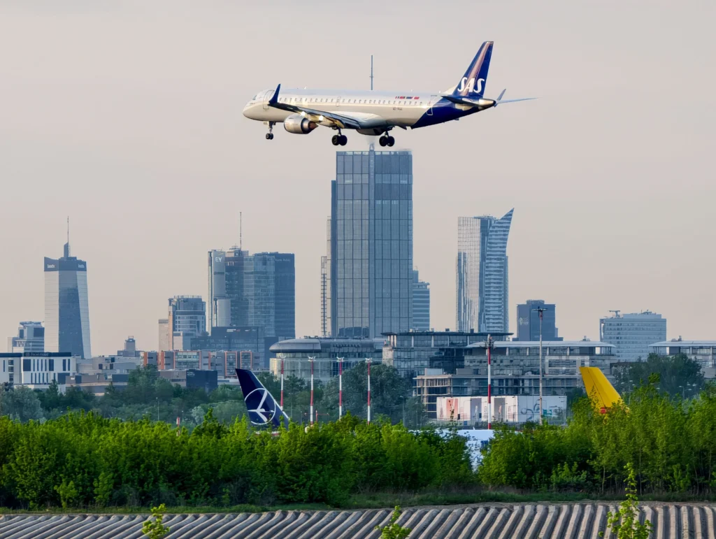 Commercial airliner (SAS) descending toward a city airport with tall glass buildings in the background. Flying over greenery and runway area.
