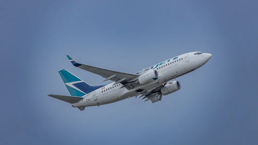 Commercial passenger jet with teal and blue tail logo flying upward in a cloudy blue sky, engines visible under wings