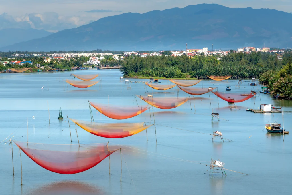 Coastal scene with orange fishing nets stretched over calm water and a town on shore, mountains in the distance