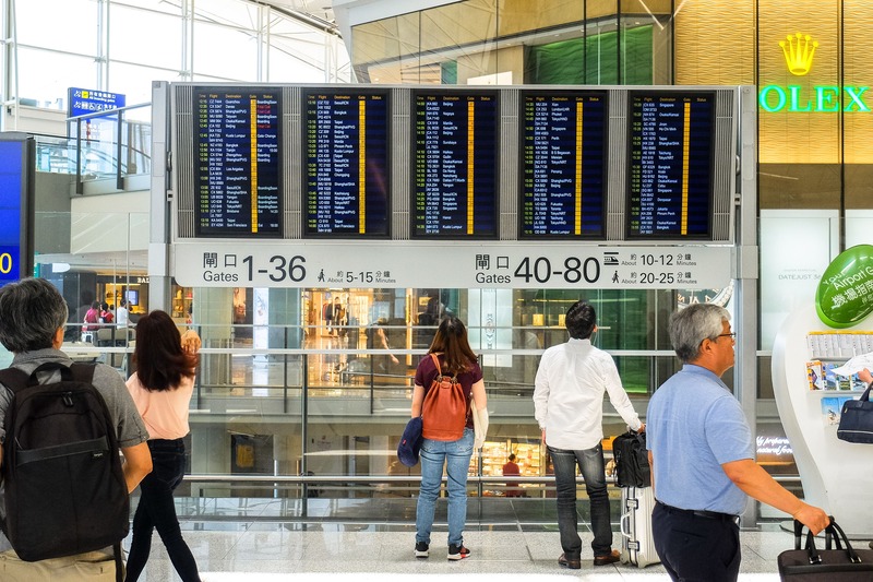 Airport departure boards showing gate numbers and flight info above a busy concourse with travelers looking at signs for Gates 1-36 and 40-80.
