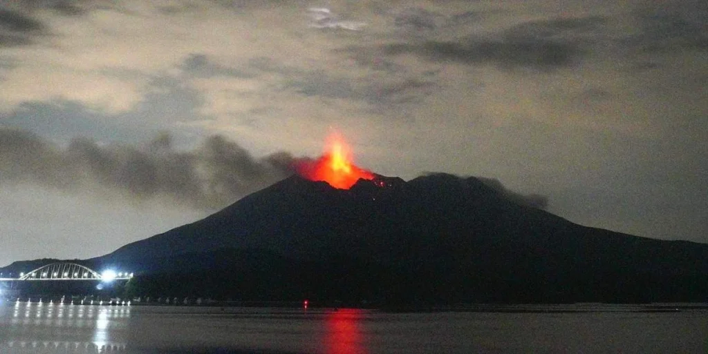 Night scene of a volcano erupting with lava plume and glow, smoke rising over a dark hill, with a lit bridge and its reflection in the water to the left.
