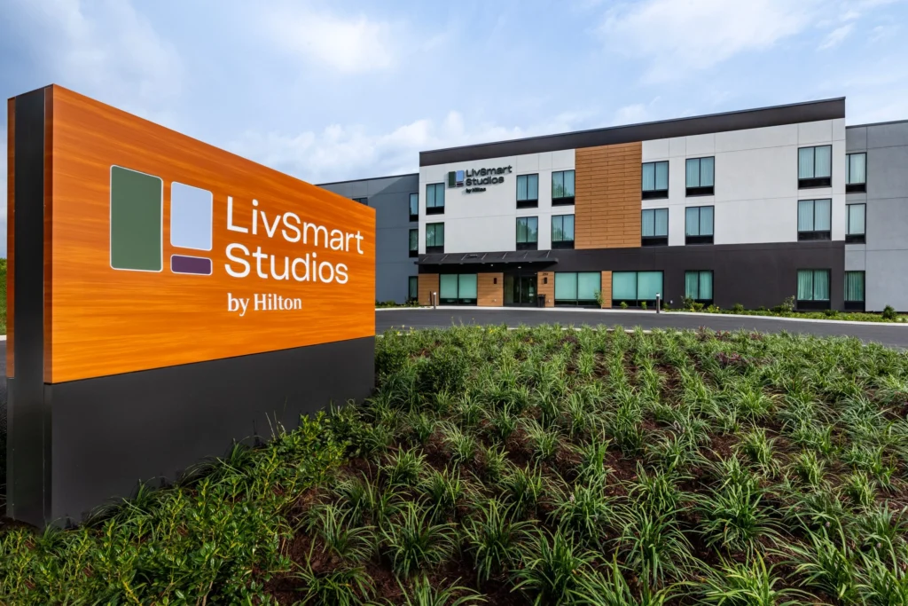Exterior view of LivSmart Studios by Hilton: an orange sign with the logo in the foreground and a modern hotel building with landscaping in the background.