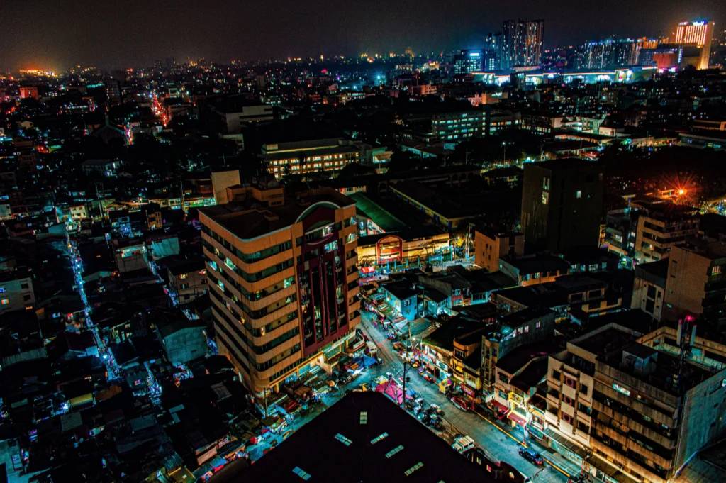 Aerial night view of a densely packed city with illuminated streets and skyline lights, seen from above across a sprawling urban area.
