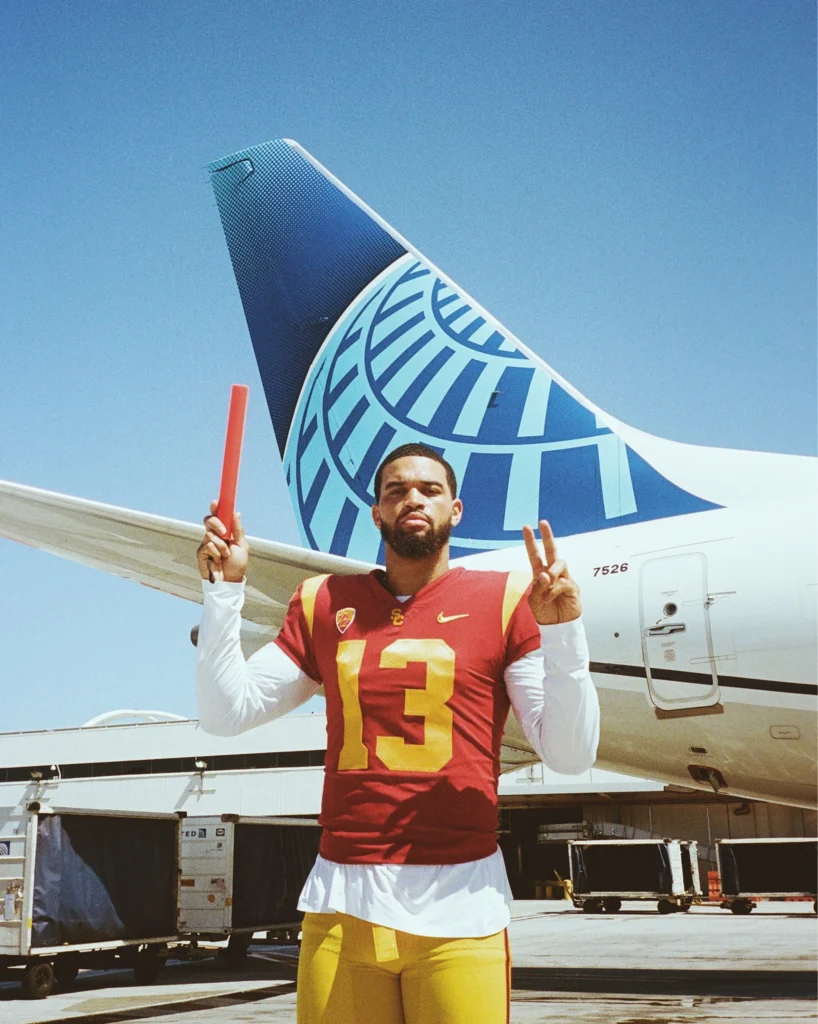 Football player in a red jersey with number 13 poses with an orange baton on the tarmac, plane tail in the background.