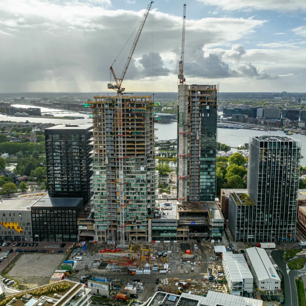 Two cranes loom over two under-construction high-rise buildings in a city by the river, with construction equipment and materials scattered on the site and surrounding modern glass buildings.