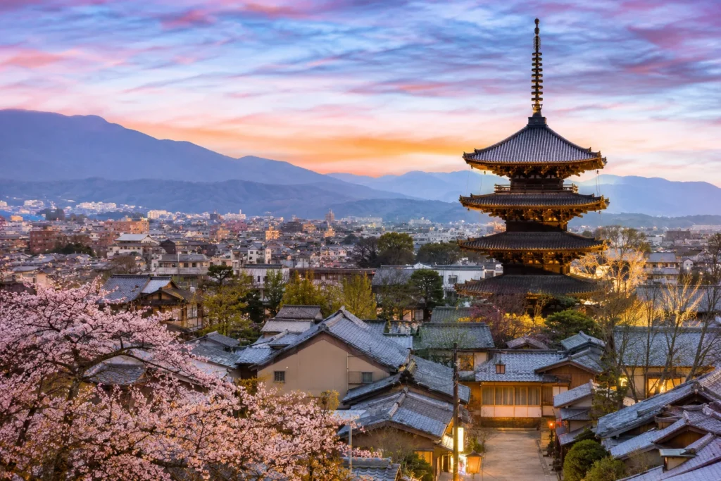 Five-story pagoda rises among traditional wooden houses as cherry blossoms bloom in the foreground at sunset, with distant mountains.