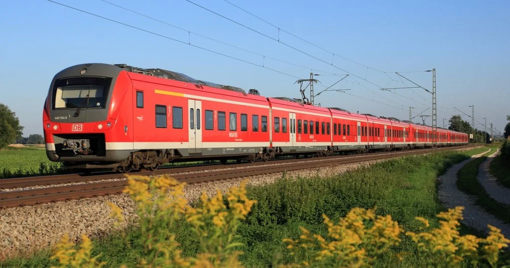 Red DB passenger train with multiple cars on a rural track, overhead power lines visible.