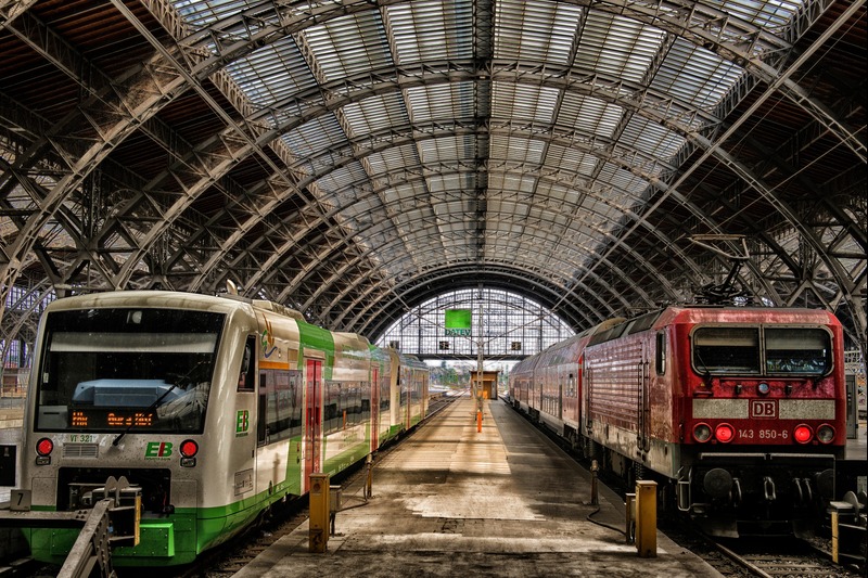 Two trains sit on parallel tracks inside a large arched train station with a glass roof. Central platform extends into the distance.