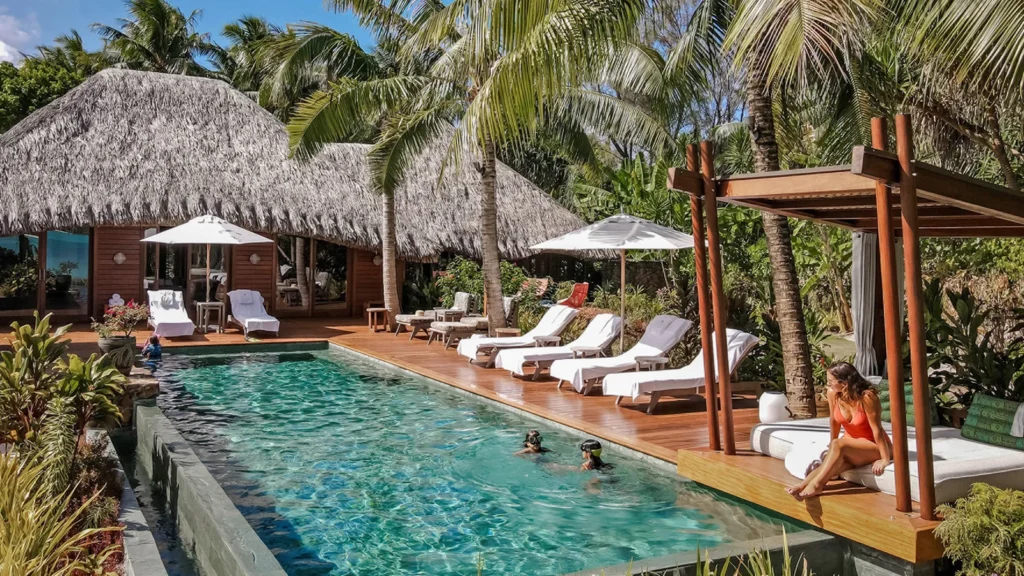 Poolside resort scene with a long rectangular pool, thatched-roof cottages, and white lounge chairs under umbrellas along the deck.