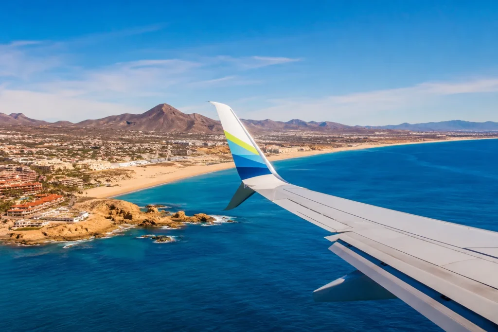 Wing of an airplane over a sunny coastal landscape: blue sea, sandy beach, rocky outcrops, and a town along the shore with mountains in the distance.