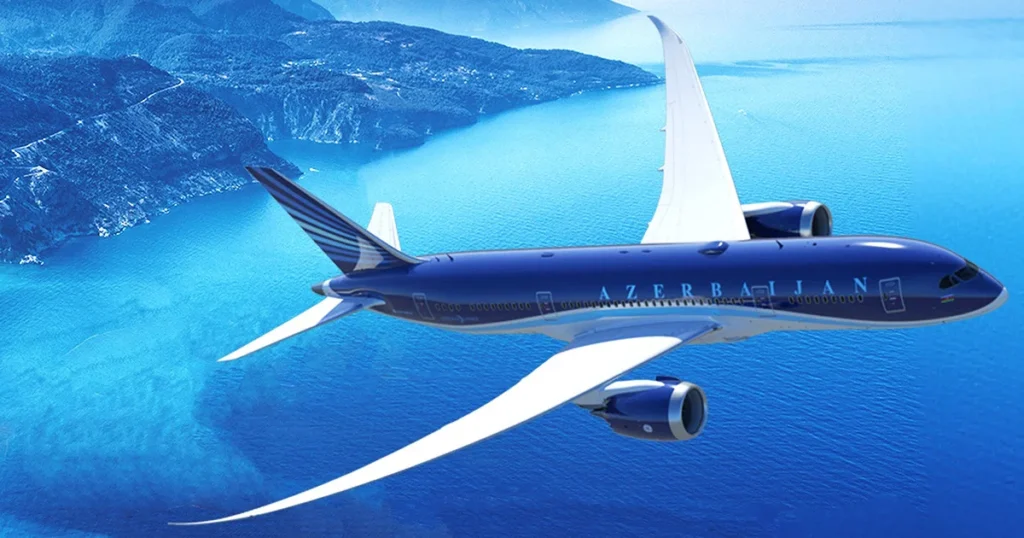 Azerbaijan Airlines jet in blue livery flies over a turquoise sea and rocky coastline below.