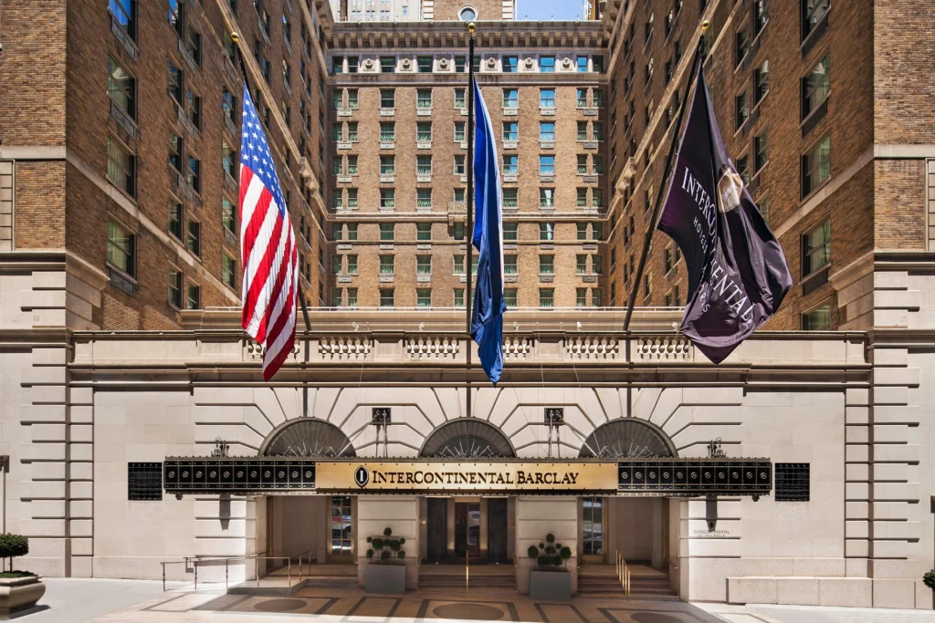 Front entrance of the InterContinental Barclay hotel with three flags (U.S., blue, and InterContinental) displayed above the doorway in a brick building courtyard.