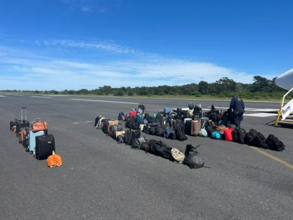 Row of suitcases and bags lined up on an airport tarmac beside a plane stairs, with a few people standing nearby under a clear blue sky