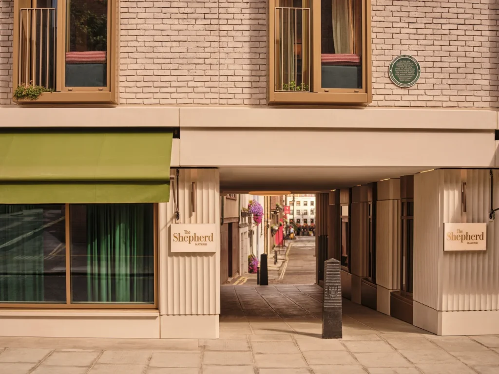 Brick building with two large windows and a green awning, opening to a pedestrian alley that leads to a street; sign reads The Shepherd Mayfair on the wall.