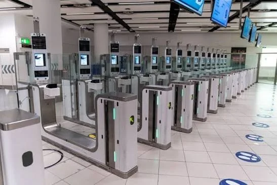 Row of airport self-check-in or security gates with touch-screen kiosks and green indicator lights on each barrier.