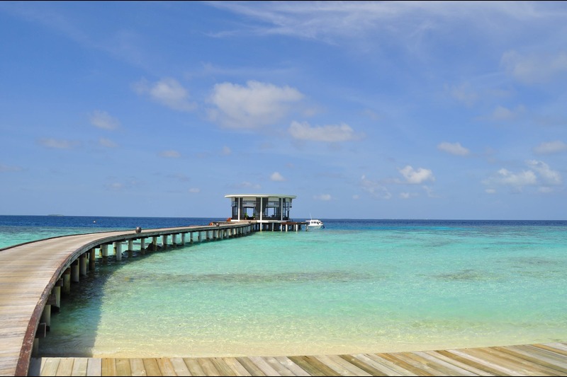 Curved wooden pier extends over turquoise water toward a small pavilion on stilts at the end, with a boat nearby.
